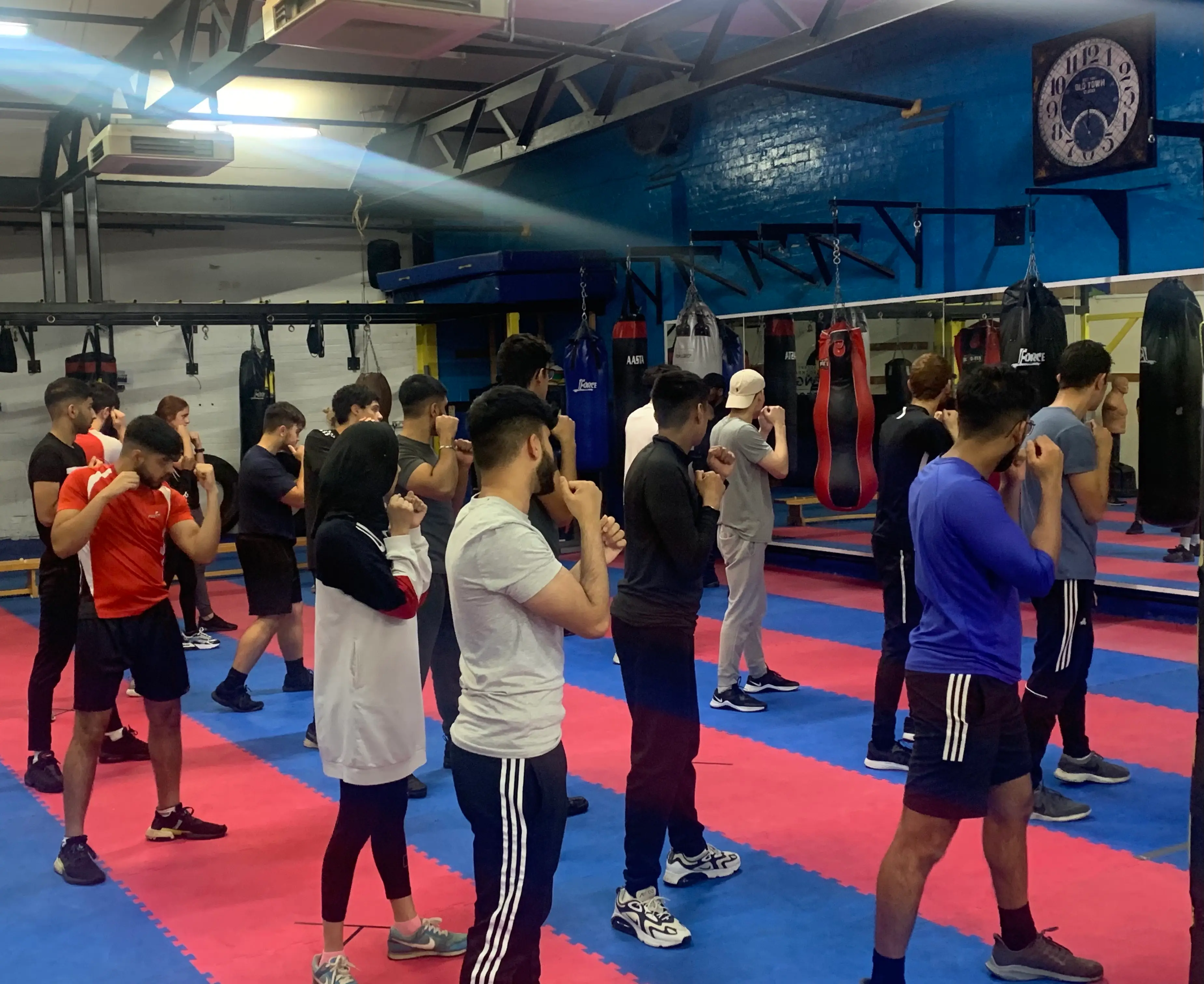 Adults standing in guard position across the gym floor during a Box session, with punch bags along the wall behind them